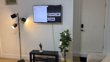 Ein Wohnzimmer mit einem an der Wand befestigten Flachbildfernseher, einer Stehlampe, einem kleinen schwarzen Tisch mit einer Pflanze und einer Tür im Hintergrund.