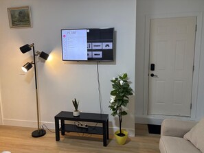 Ein Wohnzimmer mit einem an der Wand befestigten Flachbildfernseher, einer Stehlampe, einem kleinen schwarzen Tisch mit einer Pflanze und einer Tür im Hintergrund.