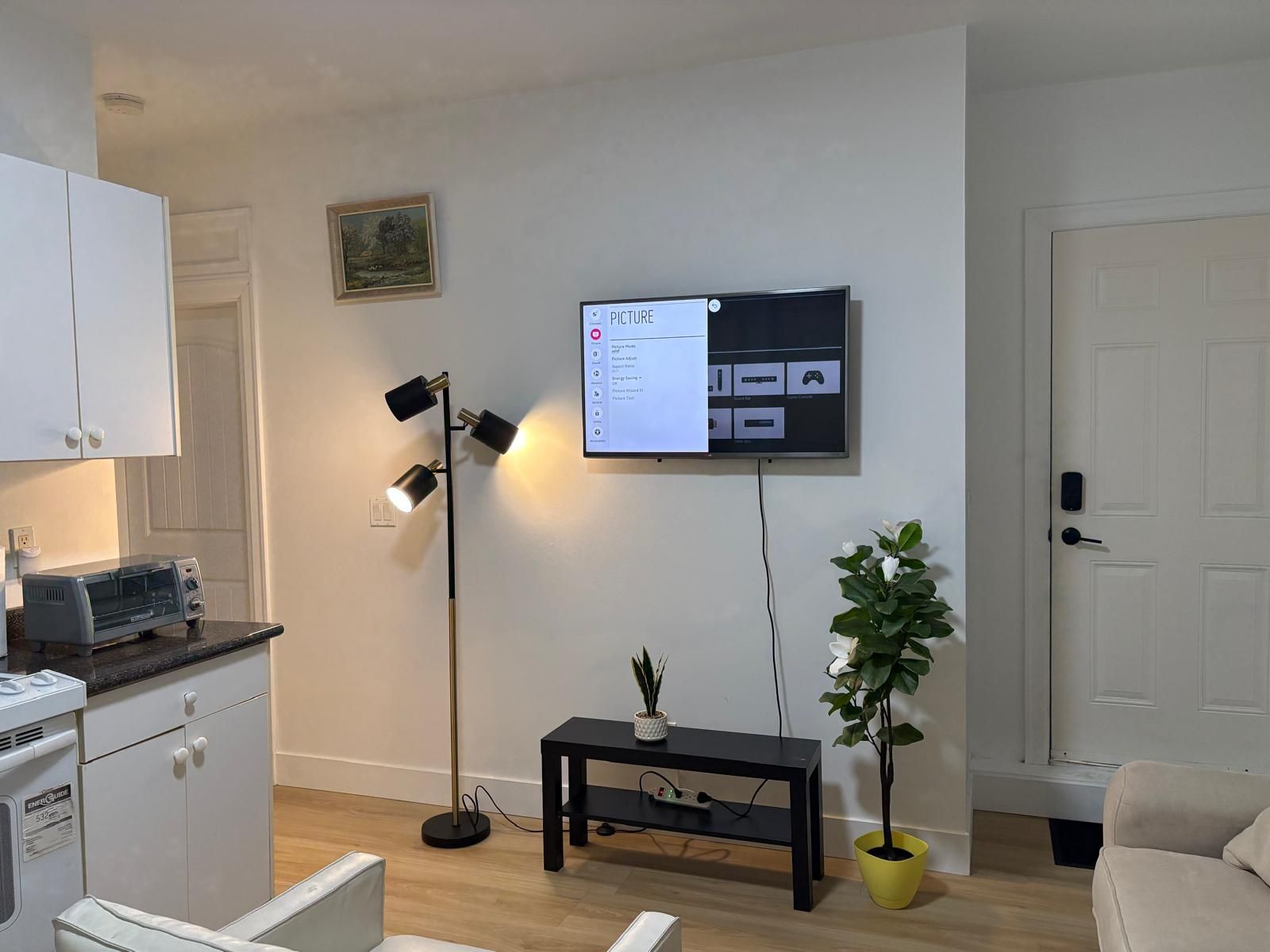 A living room with a flat-screen TV mounted on the wall, a floor lamp, a small table with a plant, and a sofa.