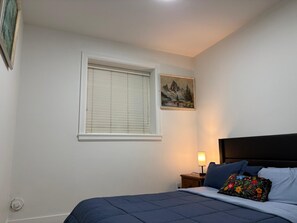 A bedroom with a bed, a nightstand, a lamp, a window with blinds, and a painting on the wall.