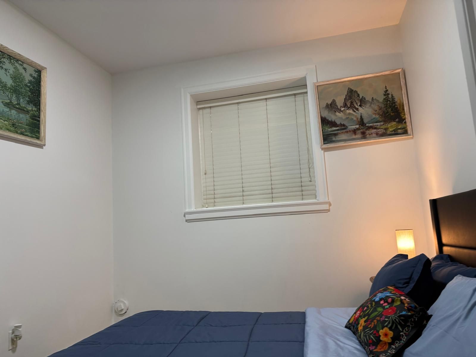 Une chambre à coucher avec un lit, une fenêtre avec des stores et deux tableaux accrochés au mur.
