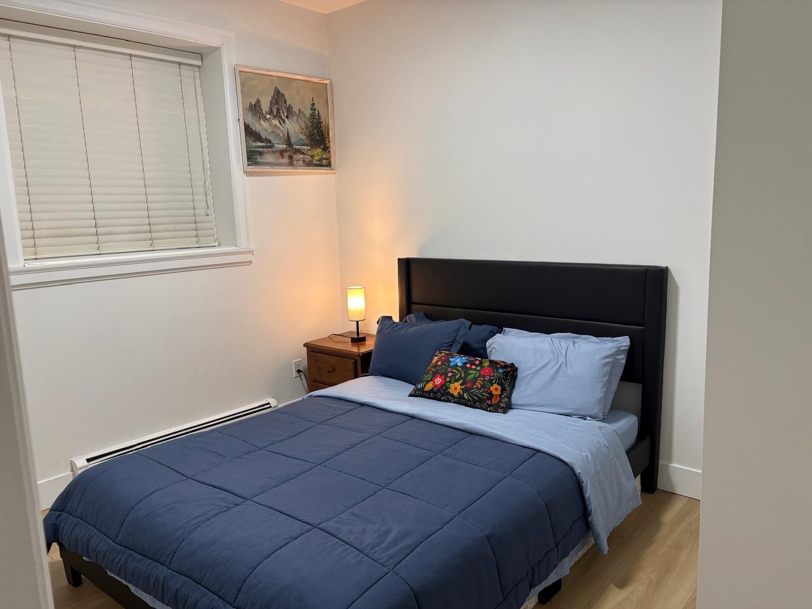 Een slaapkamer met een bed, nachtkastje, lamp en een schilderij aan de muur.