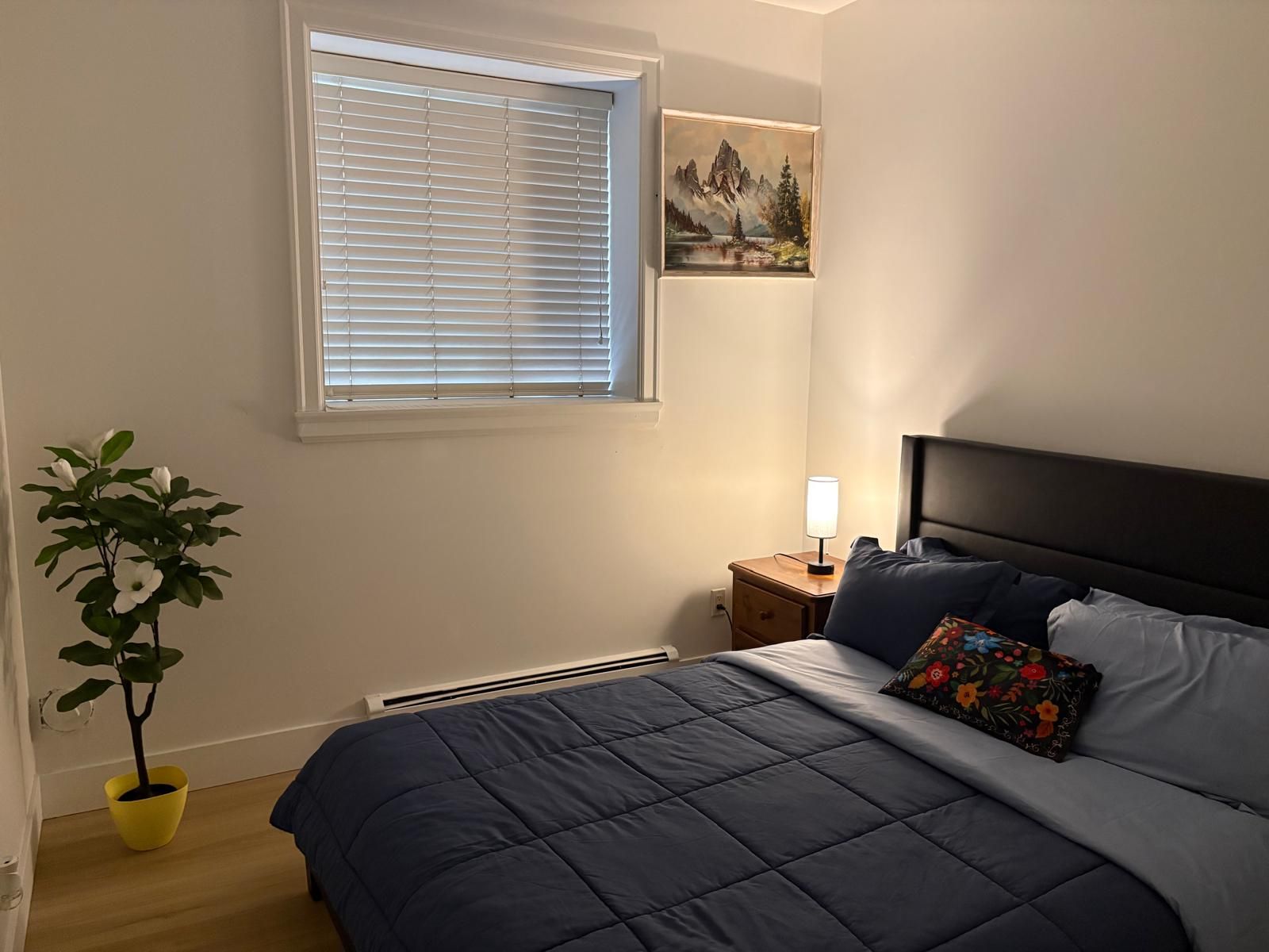 Quarto com cama, mesa de cabeceira, luminária, janela com persianas, pintura e planta em vaso.