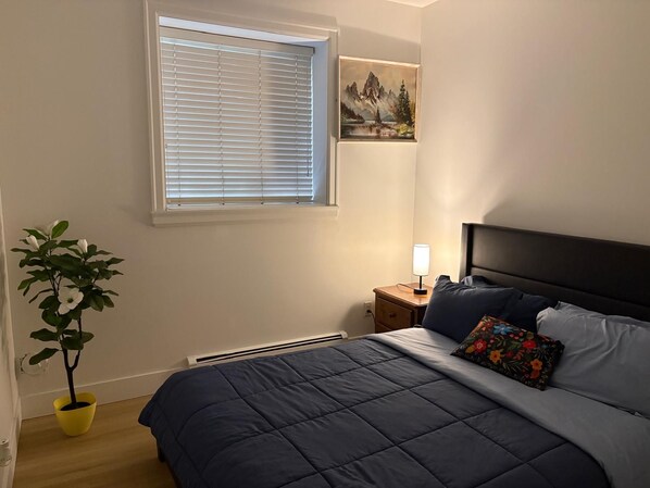 A bedroom with a bed, a nightstand, a lamp, a window with blinds, a painting, and a potted plant.