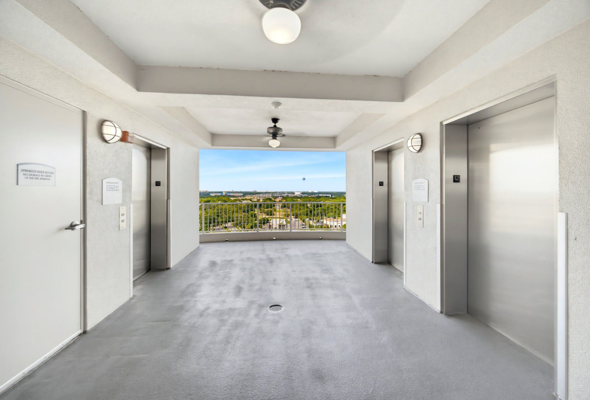 A corridor with two elevators, a ceiling fan, and a view of a cityscape through a balcony railing.