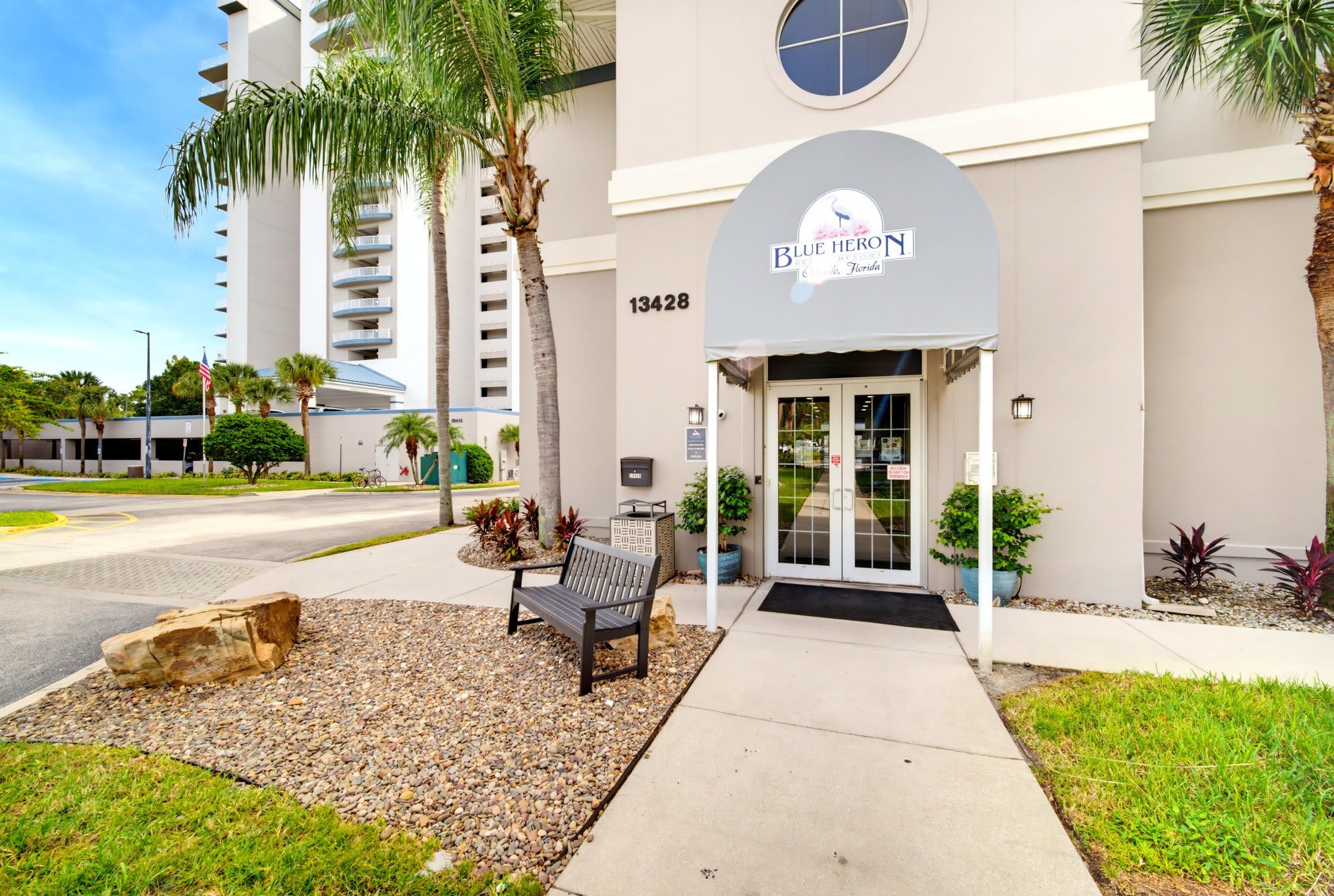 A hotel entrance with a bench, palm trees, and a clear sky.