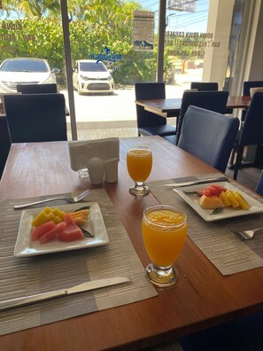 A breakfast table with plates of fruit and two glasses of orange juice, set on a wooden table with a view of parked cars and greenery outside.