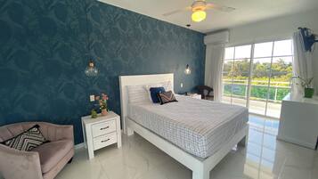 Un dormitorio con una cama grande, una mesita de noche, una silla, un ventilador de techo y un ventanal con vistas a zonas verdes.