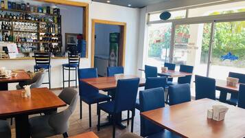 L’intérieur d’un restaurant présente de nombreuses tables et chaises, un bar avec diverses bouteilles et verres, et offre une vue sur la verdure à l’extérieur.