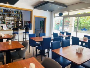 A restaurant interior with multiple tables and chairs, a bar area with various bottles and glasses, and a view of greenery outside. - Hotel Down Town Cabrera (Cabrera)
