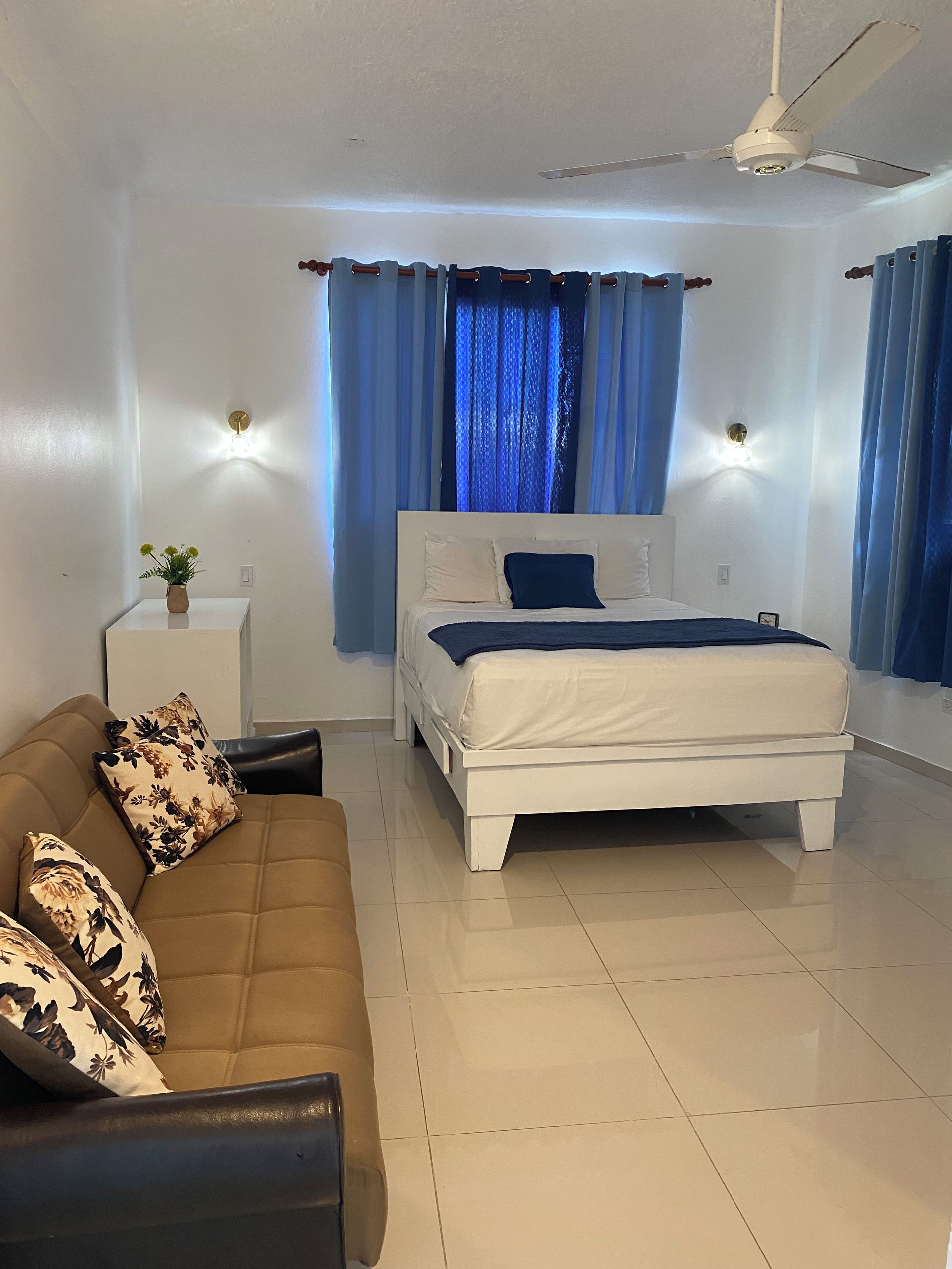 Un dormitorio con cama, sofá, una mesita con un florero y cortinas.