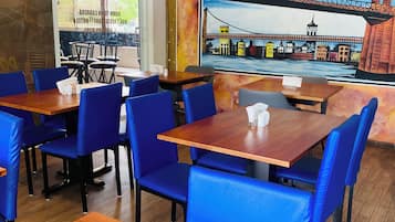 A restaurant interior with wooden tables and blue chairs, a mural of a bridge, and an air conditioning unit on the wall.
