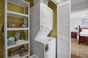 A compact laundry area with a white washer and dryer, shelves with cleaning supplies, and a bed in the adjacent room. - Amelia Island Retreat | Walk to Beach, Pool & Balcony (Fernandina Beach)