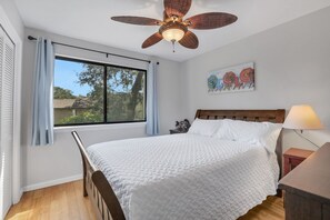 A bedroom with a bed, a ceiling fan, a window with curtains, a nightstand, a lamp, and a wall art piece. - Amelia Island Retreat | Walk to Beach, Pool & Balcony (Fernandina Beach)