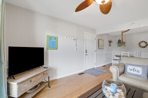 Ein Wohnzimmer mit einem Flachbildfernseher, einer Holzunterhaltungsecke, einer weißen Tür, einem weißen Sessel, einem beigen Sofa mit einem 'relax'-Kissen und einem Deckenventilator.
