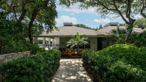 A building with a covered porch, surrounded by lush greenery and a paved pathway leading to the entrance. - SW202 - Sea Woods Retreat | Walk to Beach + Pools & Trails (New Smyrna Beach)