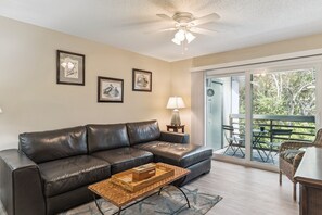 A living room with a leather sofa, a coffee table, and a ceiling fan. There are framed pictures on the wall and a view of the outdoors through a sliding glass door. - SW202 - Sea Woods Retreat | Walk to Beach + Pools & Trails (New Smyrna Beach)