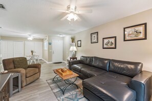 A living room with a black leather sofa, a brown armchair, a coffee table, and a ceiling fan. - SW202 - Sea Woods Retreat | Walk to Beach + Pools & Trails (New Smyrna Beach)