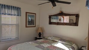 A bedroom with a bed, a ceiling fan, a bedside lamp, a window with blinds, a framed picture, and a mirror with a rustic frame.