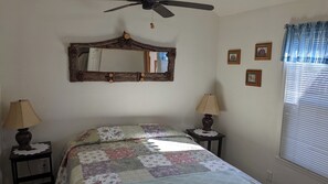 A bedroom with a bed, two bedside lamps, a ceiling fan, a mirror, and a window with blinds. - The Apple Place Room #3 (Miramonte)