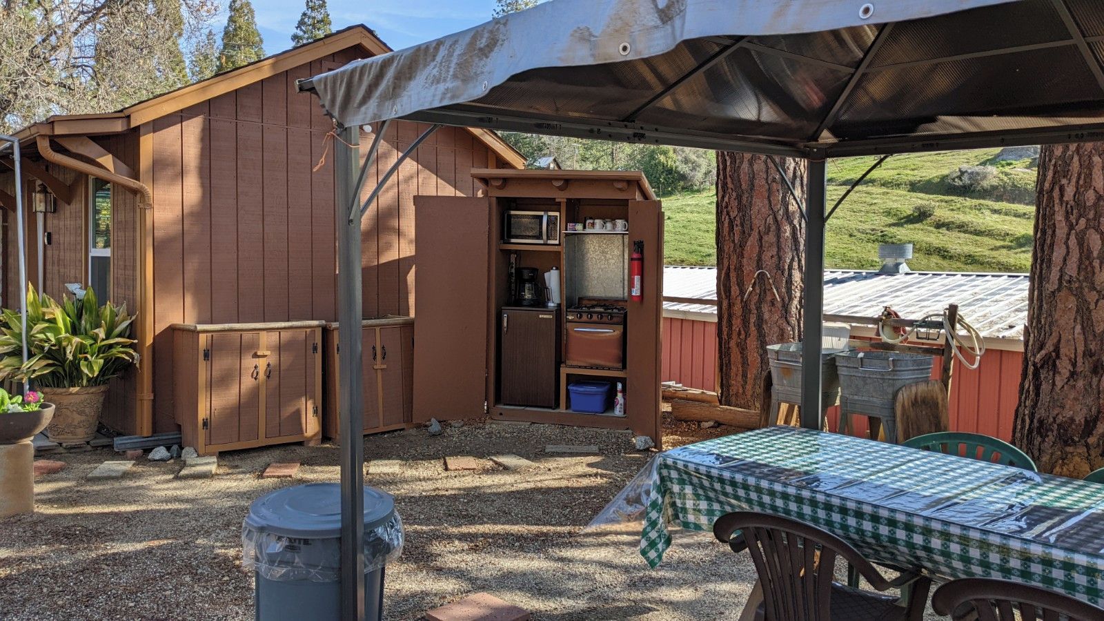 A rustic outdoor kitchen area with a table and chairs, a wooden structure with a door, and a metal canopy.