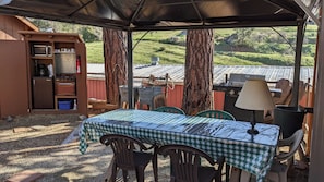 Un área de comedor al aire libre techada con una mesa para dos, rodeada de sillas, una lámpara y una parrilla.