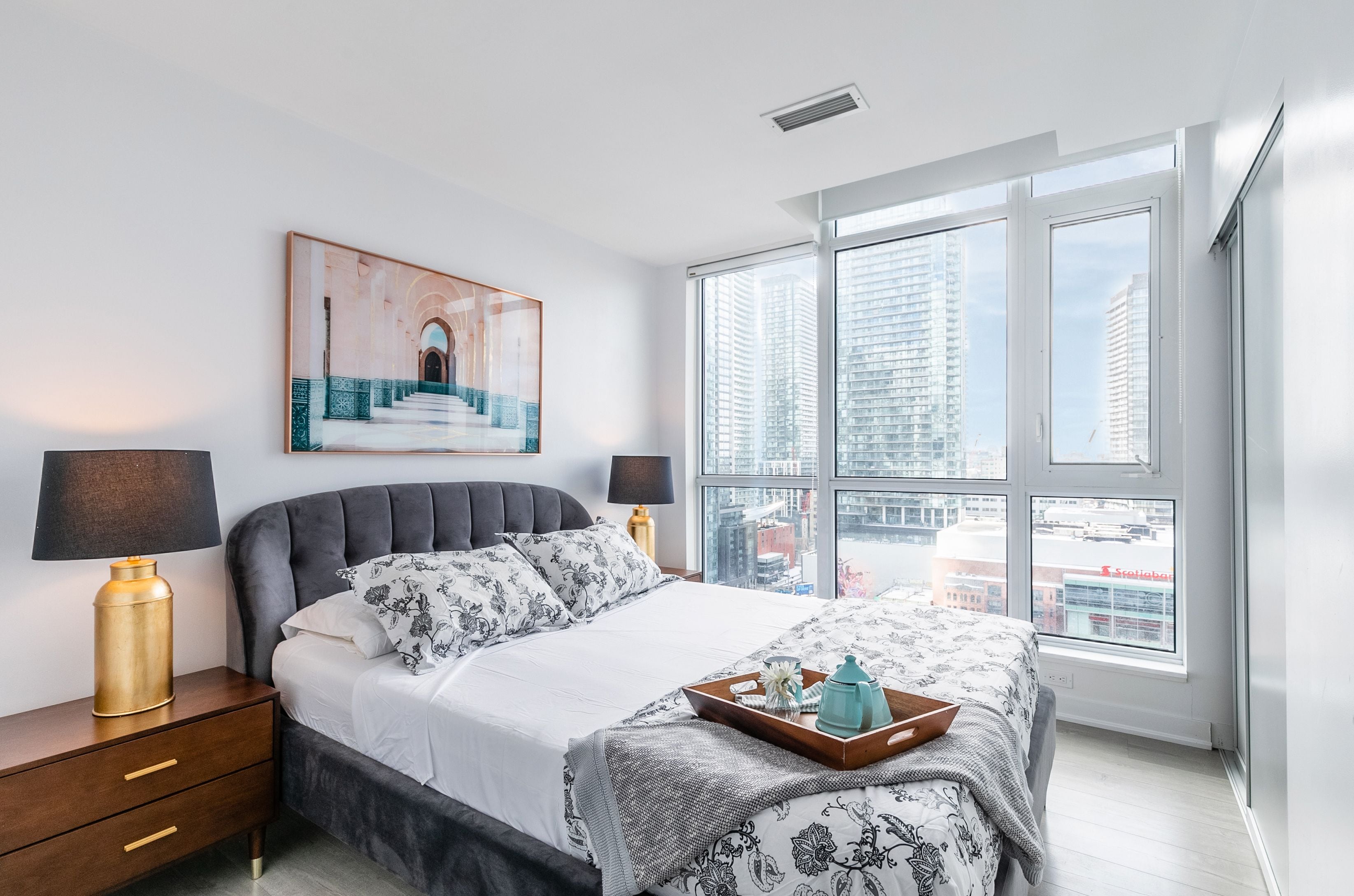 A modern bedroom with a large bed, two bedside lamps, a framed artwork, and a city view through large windows.