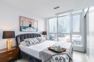Un dormitorio moderno con una cama grande, dos lámparas de noche, una obra de arte enmarcada y vistas de la ciudad a través de amplias ventanas.