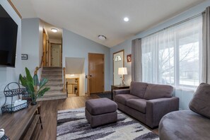 A living room with a sofa, ottoman, and a TV. There's a staircase leading to another room, and a window with curtains. - Your Home Away From Home! Only 20-25 Minutes to Madison! (Stoughton)