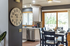 Una cocina moderna con un reloj de madera, un comedor con una mesa lista para dos personas, y vistas al exterior a través de amplios ventanales.