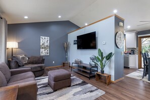 A modern living room with a flat-screen TV, a sofa, a coffee table, and a dining area with a kitchen visible in the background. - Your Home Away From Home! Only 20-25 Minutes to Madison! (Stoughton)