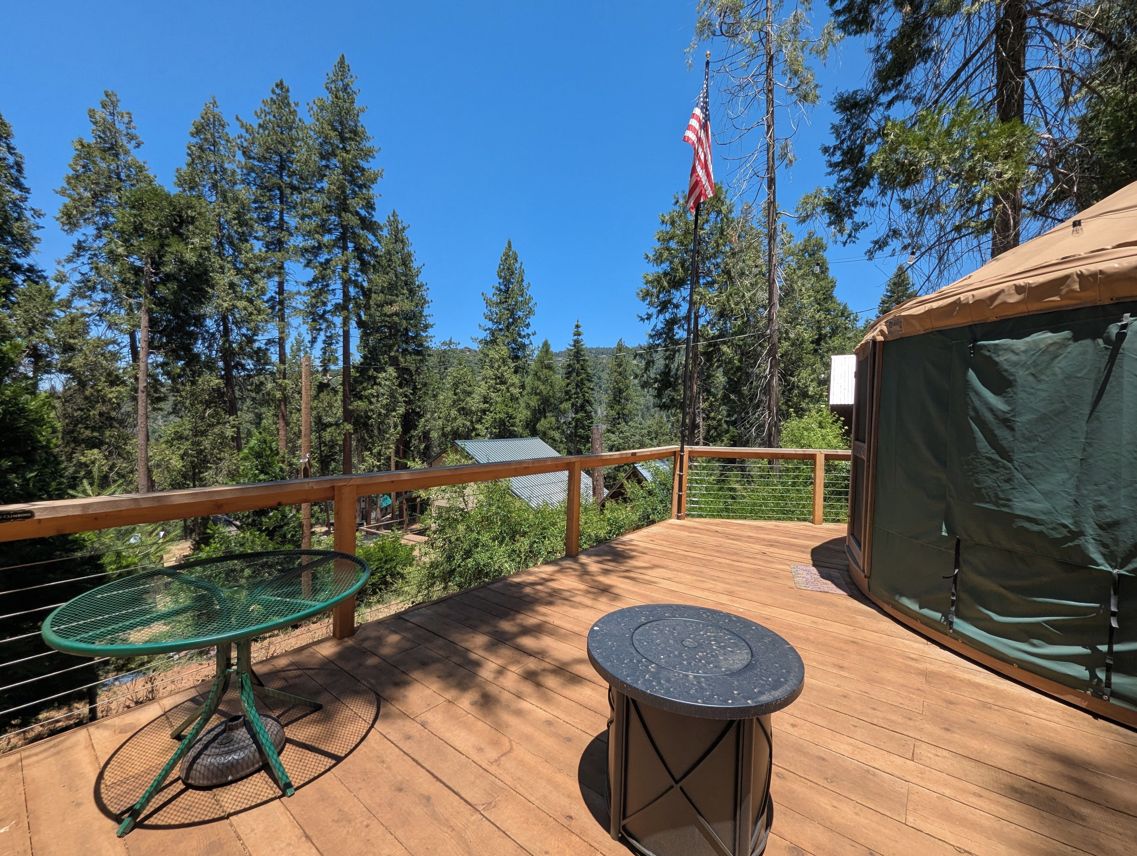 A wooden deck with a metal table and a lantern, surrounded by trees and a tent.