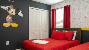 Ein Hotelzimmer mit Wandsticker von Mickey Mouse, einem Bett mit roter Bettwäsche, einem weißen Kleiderschrank und einem Fenster mit roten Vorhängen.