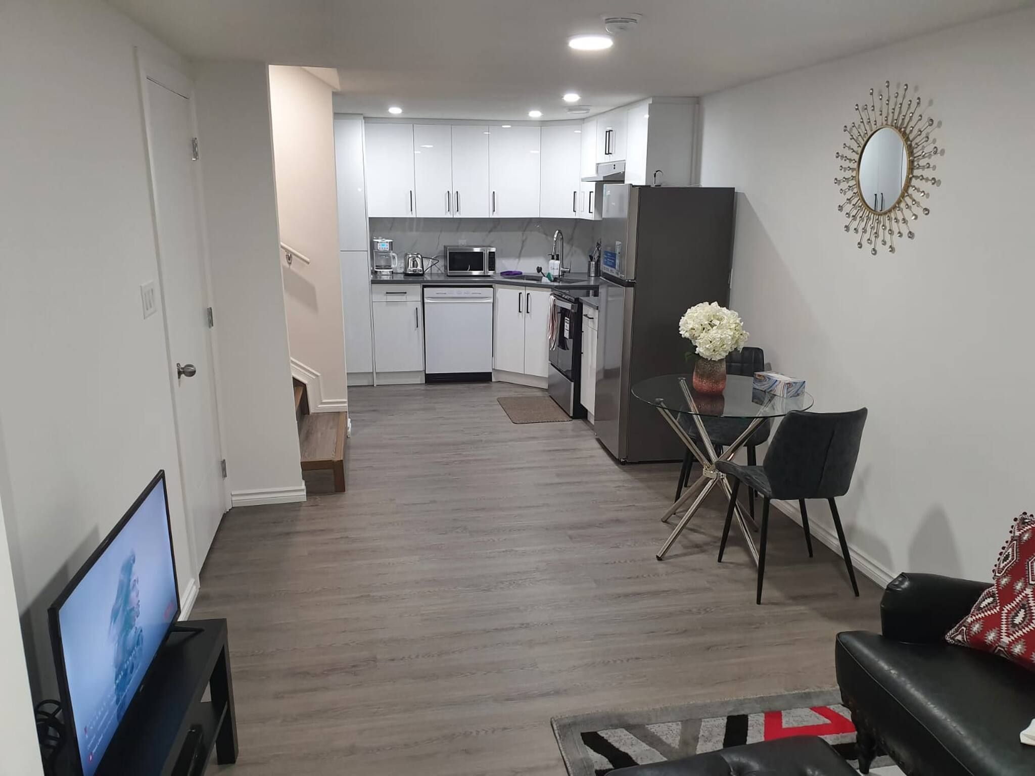 A modern kitchen with white cabinets, a dining area with a glass table, and a living room with a black sofa.