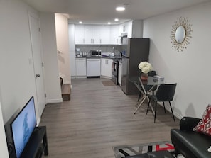 A modern kitchen with white cabinets, a dining area with a glass table, and a living room with a black sofa. - Beautiful Tranquil 1bedroom Basement Suite (Beaumont)