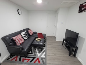 A living room with a black leather sofa, red and white patterned cushions, a black ottoman, a television on a stand, and a patterned rug.