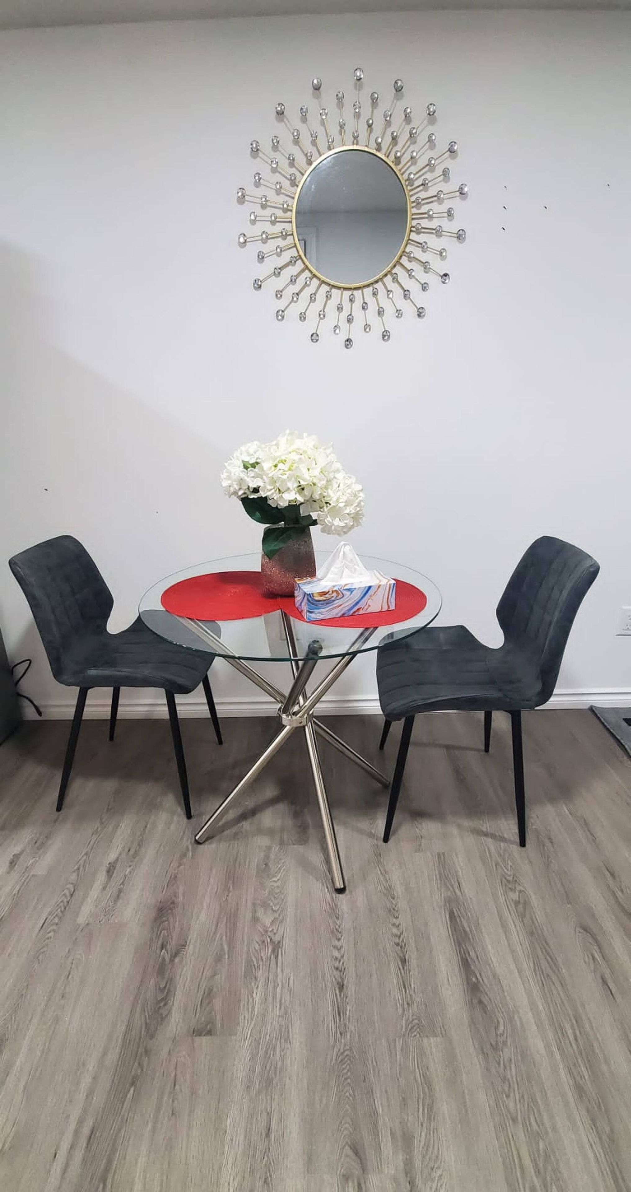 Un comedor con una mesa redonda, dos sillas, un florero con flores y un espejo en la pared.