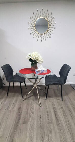 Un comedor con una mesa redonda, dos sillas, un florero con flores y un espejo en la pared.