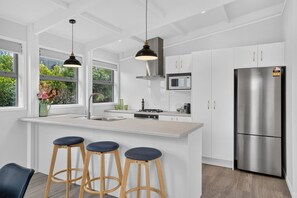 A modern kitchen with white cabinetry, a stainless steel refrigerator, and a bar area with stools. - Blue Moon Coastal Escape (Langs Beach)