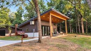 A modern house with a covered patio, a barbecue grill, and a large window. - Texas Tiny Cabins #1 - Cozy Getaway Near Texoma (Grayson County)