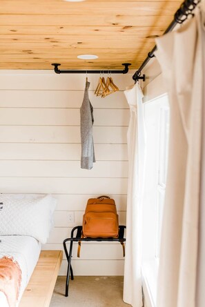 A bedroom with a bed, a hanging rack with clothes, a suitcase on a chair, and a window with curtains. - Texas Tiny Cabins #1 - Cozy Getaway Near Texoma (Grayson County)