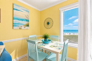 A dining area with a table and chairs, a painting, a decorative wall piece, and a view of the beach through a window. - A Place at the Beach - Windy Hill 308 - 2BR 2BA Condo North Myrtle Beach Pool & Hot tub (North Myrtle Beach)