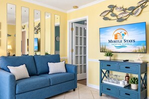 Quarto com sofá azul, parede espelhada, televisão exibindo um anúncio de aluguel por temporada e um painel de parede decorativo com tema de peixes.