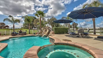 Une piscine avec un jacuzzi, entourée de mobilier d’extérieur et de parasols, le tout se fondant dans un décor de palmiers et d’un ciel dégagé.
