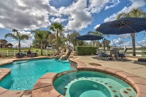 Une piscine avec un jacuzzi, entourée de mobilier d’extérieur et de parasols, le tout se fondant dans un décor de palmiers et d’un ciel dégagé.