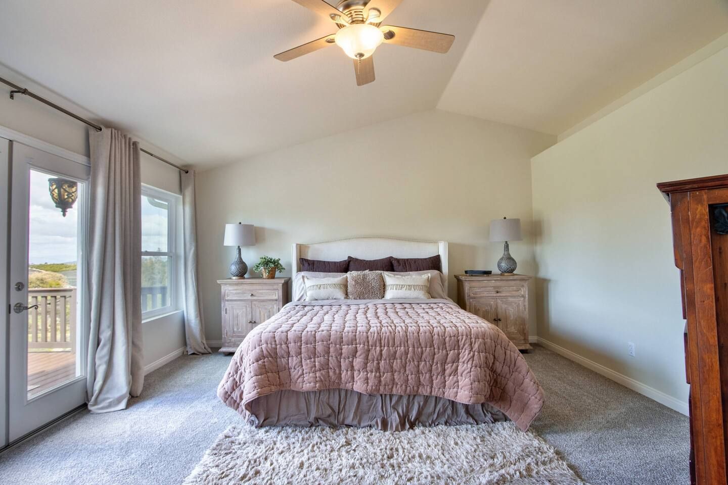 Une chambre à coucher comprenant un lit, deux tables de chevet, un ventilateur de plafond, une grande fenêtre avec des rideaux et une vue sur l’extérieur.