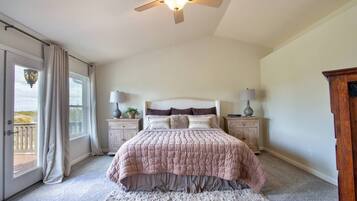 Une chambre à coucher comprenant un lit, deux tables de chevet, un ventilateur de plafond, une grande fenêtre avec des rideaux et une vue sur l’extérieur.