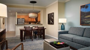 Una sala moderna con un sofá gris, una mesa de comedor de madera y sillas. Se ve una cocina al fondo, con armarios de madera y un refrigerador de acero inoxidable.