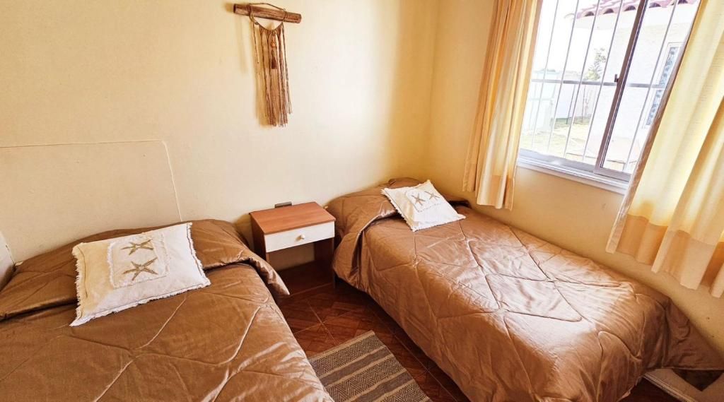 A room with two beds, a small wooden nightstand, a wall-mounted macrame decoration, and a window with curtains.