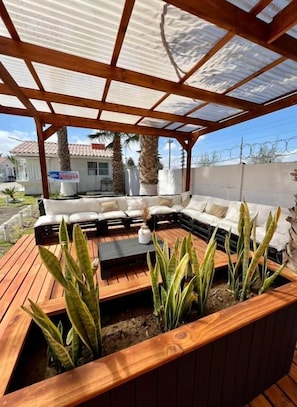 Un área de estar exterior con terraza de madera, sofá acolchado y plantas en macetas.